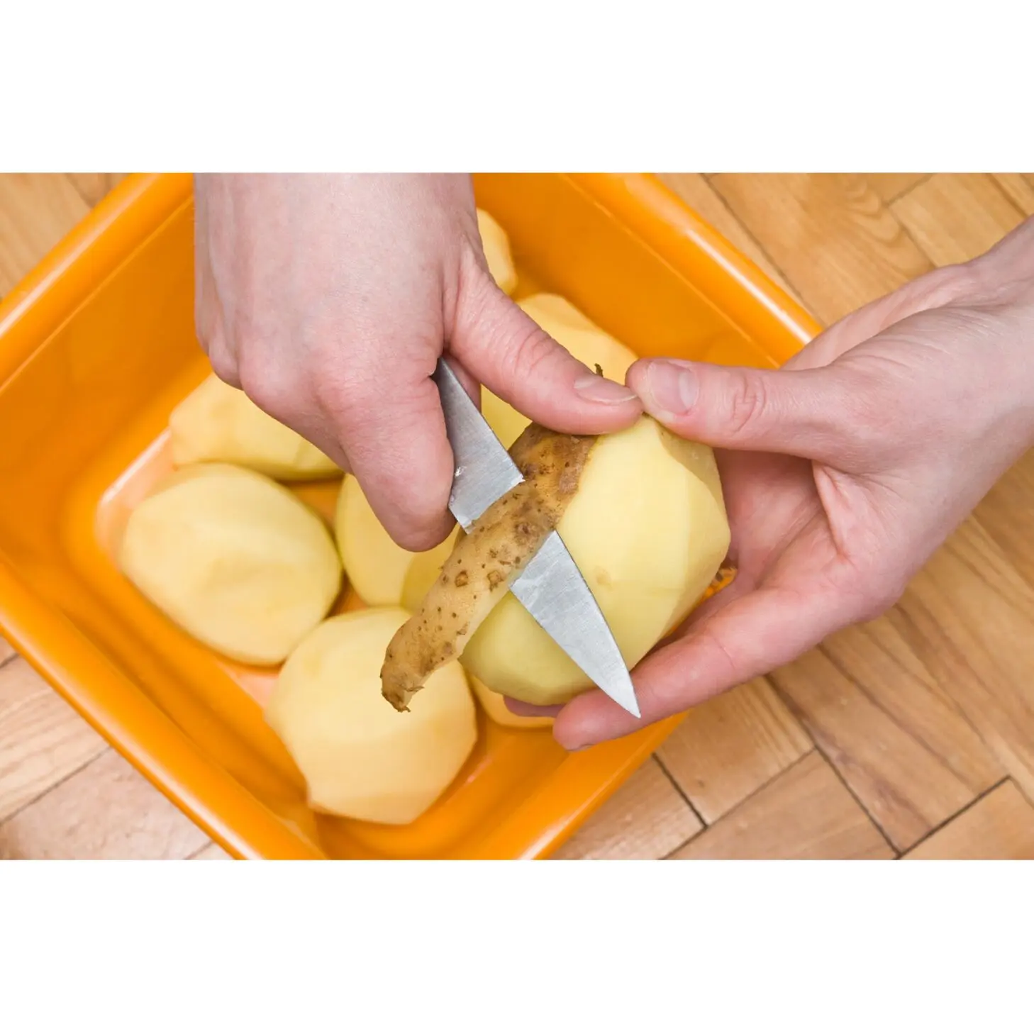 A person peeling a potato with a knife over an orange container.
